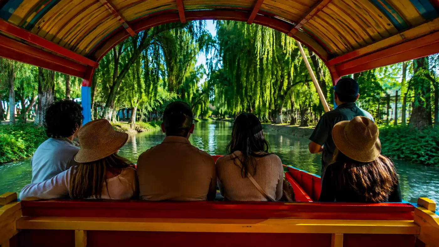 Passengers sitting along the bench of a trajinera, looking out through the painted arched canopy at willows leaning over the canal