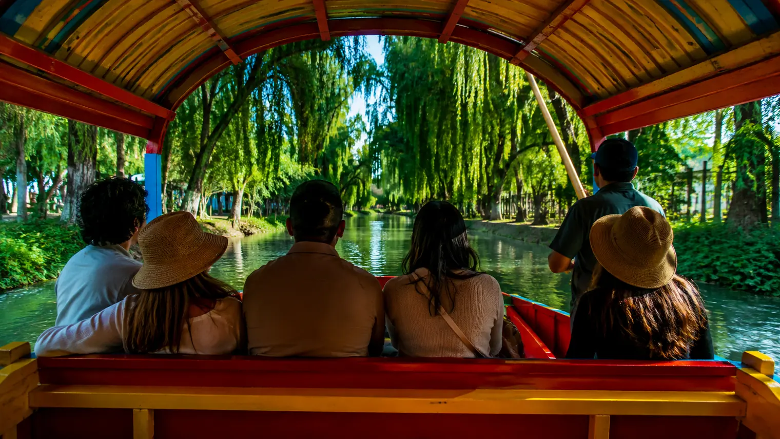 Passengers along the bench of a painted trajinera under its canopy, seen from behind, looking out at the water and the willows