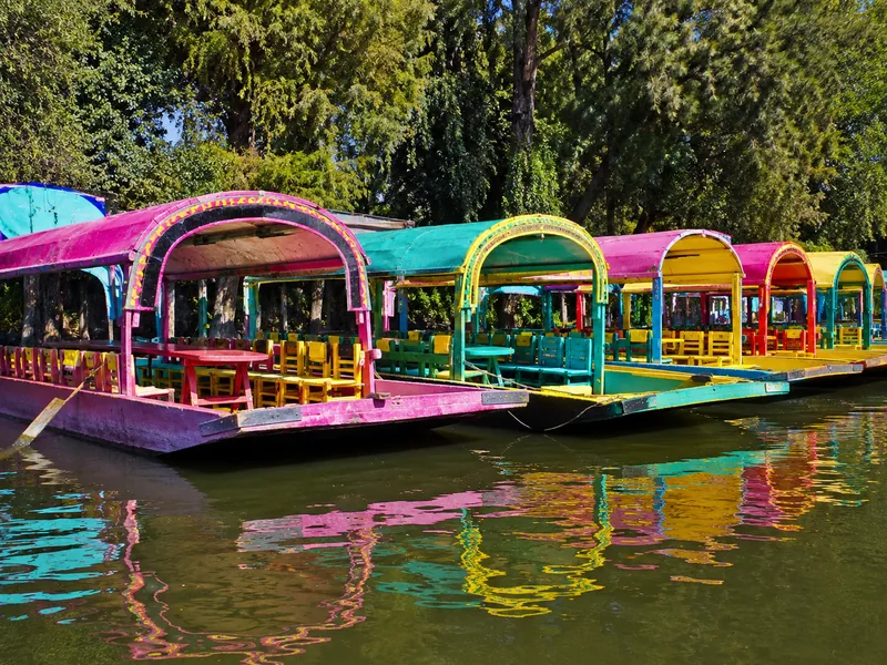 Trajineras moored in a row, seen from the waterline, arched canopies painted pink, turquoise and yellow above green canal water